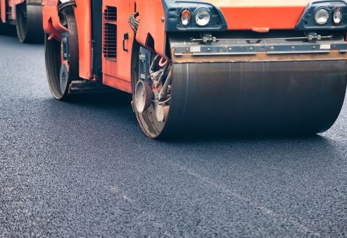 Heavy machines work diligently on a city street, laying fresh asphalt. The bright sun shines down as crews upgrade the road surface, ensuring smooth travel for vehicles and pedestrians alike.