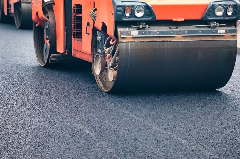 Heavy machines work diligently on a city street, laying fresh asphalt. The bright sun shines down as crews upgrade the road surface, ensuring smooth travel for vehicles and pedestrians alike.