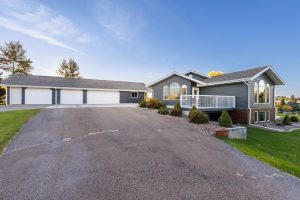 A two-story, white house with a red door situated on a grassy area beside a paved road, surrounded by trees